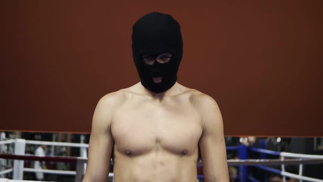 Full length footage boxer or fighter topless standing in the thief, rubber mask on the boxing ring in the gym. Caucasian with muscular torso, wearing black pants standing with black gun in a hand