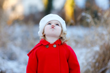 Outdoor winter portrait of little cute toddler girl in red coat and white fashion hat barret. Healthy happy baby child walking in the park on cold day with snow and snowfall. Stylish clothes for kids.