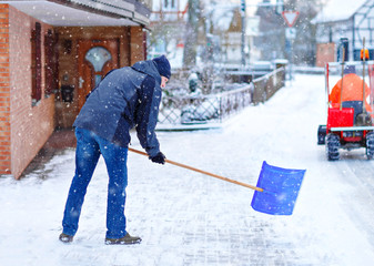 Man with snow shovel cleans sidewalks in winter during snowfall. Winter time in Europe. Young man in warm winter clothes. Snow and weather chaos in Germany. Snowstorm and heavy snowing. Schneechaos