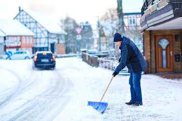 Man with snow shovel cleans sidewalks in winter during snowfall. Winter time in Europe. Young man in warm winter clothes. Snow and weather chaos in Germany. Snowstorm and heavy snowing. Schneechaos © Irina Schmidt