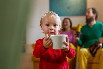 Adorable infant boy holding a big coffee mug with parents at the background.