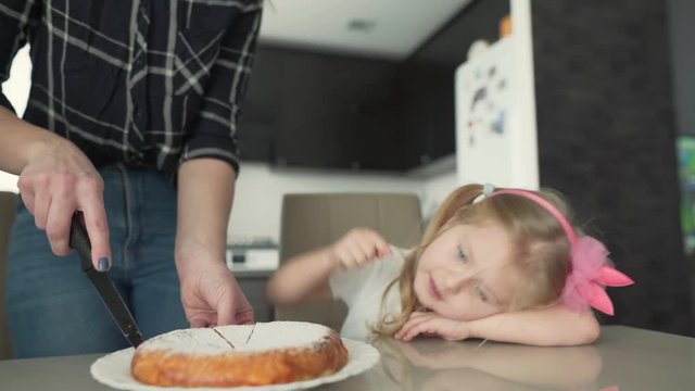 The mother cuts the pie into small pieces. The baby girl looks forward to eat a piece of pie and eats little pinches of powdered sugar from the side of the pie. The background is a bit blurred.