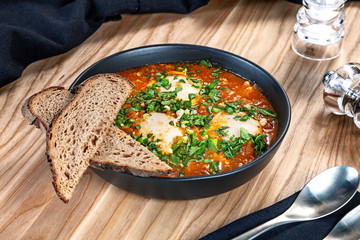 Close up view on Shakshouka, Shakshuka with green onion and bread in a black bowl and wooden background. Healthy food for menu. Free copy space. traditional Israeli cuisine
