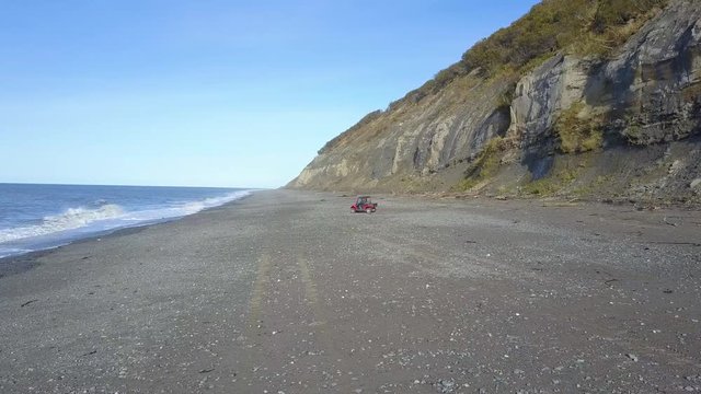 Scenic aerial drone tracking shot of ATV or All Terrain Vehicle going on beach shore next to the crashing waves and the cliffs in Deep Creek State Park in Ninilchik, Alaska