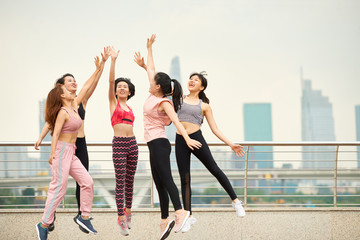 Fototapeta premium Group of laughing Asian woman in sportswear jumping and giving high five against cityscape 