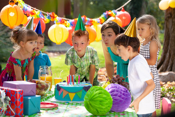 Redhead boy, his mother and friends blowing candles on birthday cake during garden party for...
