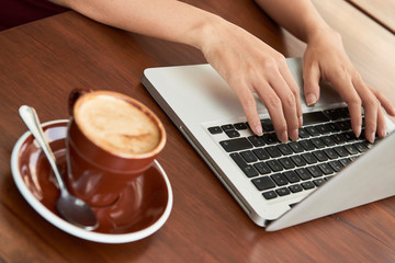 Close-up of female hands typing on keyboard of a laptop with a cup of coffee on wooden table