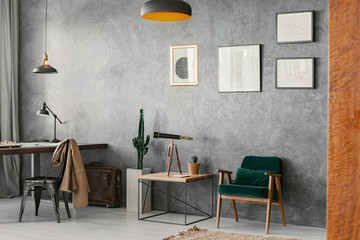Posters on concrete wall above armchair and table in home office interior with chair at desk. Real photo