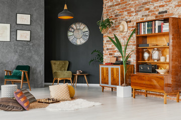 Plant next to wooden cabinet in vintage grey loft interior with pillows, posters and armchairs. Real photo