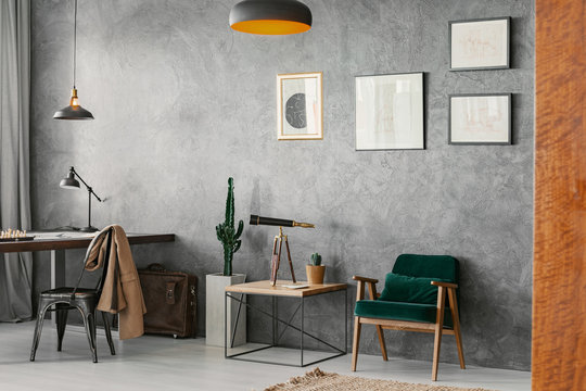 Posters On Concrete Wall Above Armchair And Table In Home Office Interior With Chair At Desk. Real Photo