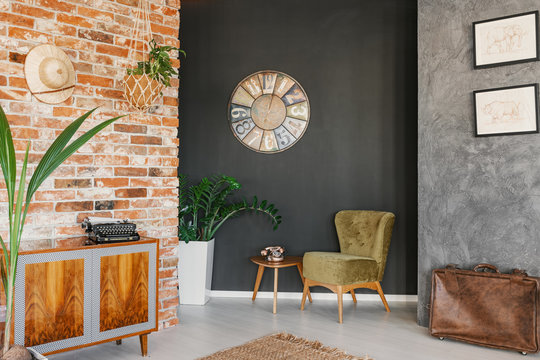Clock above table and green armchair in vintage flat interior with wooden cabinet and plants. Real photo