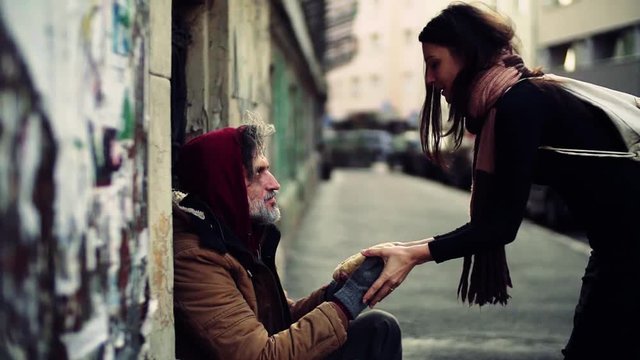 Young kind woman giving food to homeless beggar man sitting in city.