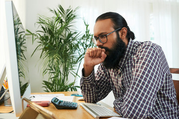 Tense young programmer in casual shirt and eyeglasses sitting by desk in front of compter monitor...