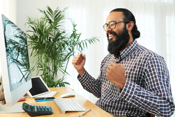 Young cheerful bearded businessman expressing gladness while siting by desk in front of computer monitor
