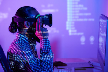 Modern programmer in vr headset sitting in front of computer monitor while coded data reflecting on him