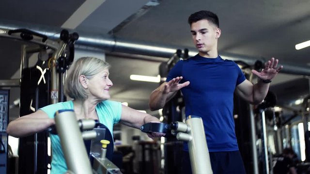A female senior with a young trainer doing strength workout exercise in gym.