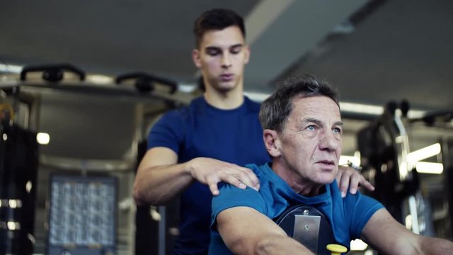A senior man with a young trainer doing strength workout exercise in gym.