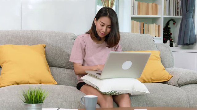 attractive young Asian woman happy working with notebook computer sitting on sofa in beautigul decoration living room, happy lifestyle concept,RAW,UHD,4K,23.98,Prores444