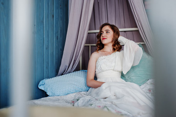 Pretty young curly gilrl in white dress sitting on bed.