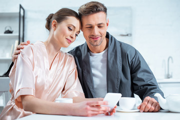 beautiful smiling couple in robes using smartphone while having tea during breakfast in kitchen