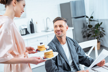 woman holding plates with pancakes while smiling man reading newspaper during breakfast in kitchen