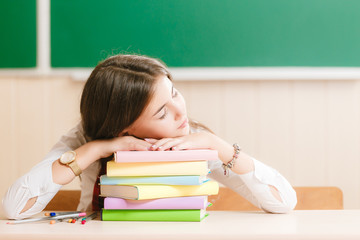 Cute brunette schoolgirl sitting at a school desk with books on the background of the blackboard