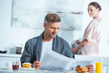 man reading newspaper during breakfast while woman cooking on background