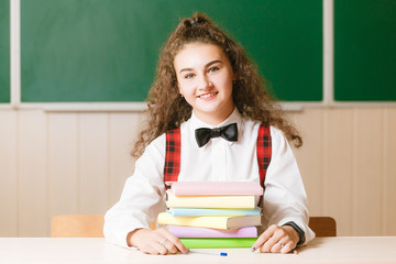 Cute brunette schoolgirl sitting at a school desk with books on the background of the blackboard