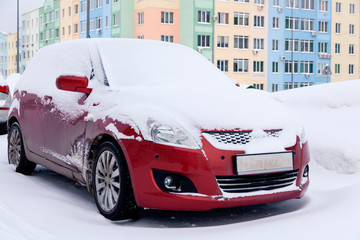 Closeup of small red dirty car covered with snow stands on background colored house. Front view. Concept snowy weather, snowfall, bad northern weather conditions, low battery, severe frost