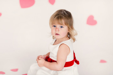 Obraz na płótnie Canvas Little blonde girl in white dress with red ribbon on the white floor with pink hearts on the St. Valentine's day. Mother's woman day concept