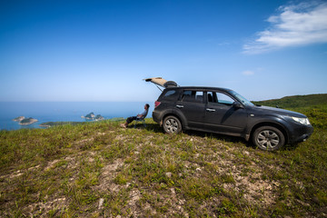 Fototapeta na wymiar Man sitting at car's shadow at the top of the hill at sea coast
