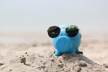 Obraz premium Blue piggy bank with sunglasses on sea beach, Saving planning for Travel budget of holiday concept
