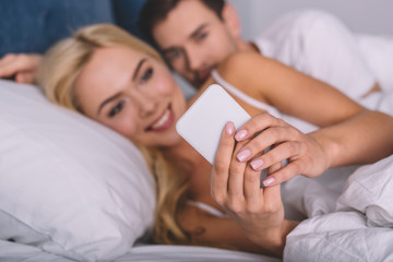 Obraz na płótnie Canvas selective focus of young man peeking at smiling girlfriend using smartphone in bed, distrust concept