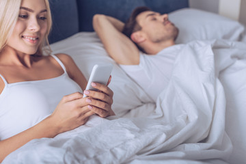 Obraz na płótnie Canvas beautiful smiling young woman using smartphone while lying in bed with sleeping husband