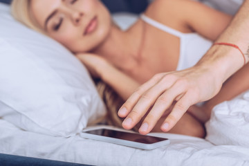 cropped shot of man reaching for smartphone while young wife sleeping in bed, distrust concept
