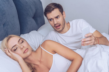 angry young man holding smartphone and looking at sleeping girlfriend in bed