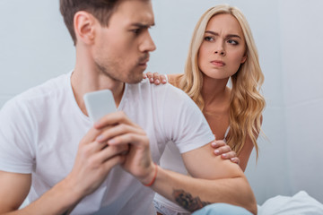 angry young woman looking at boyfriend using smartphone in bedroom, relationship problem concept