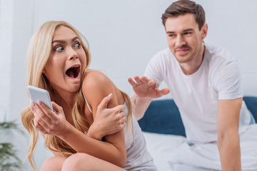 husband reaching to scared young wife using smartphone on bed, mistrust concept