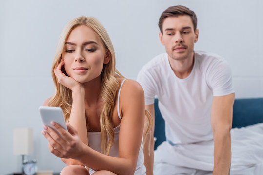 smiling young woman using smartphone and jealous man peeking behind on bed