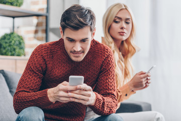 jealous young woman with smartphone looking at smiling boyfriend using smartphone at home, relationship problem concept - obrazy, fototapety, plakaty