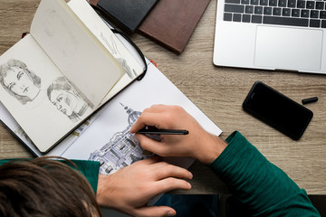 overhead back view of man drawing in album on wooden table next to laptop and smartphone - Powered by Adobe