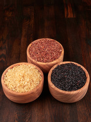 Uncooked black, red and integral rice in olive wood bowls, on a dark rustic wooden board.