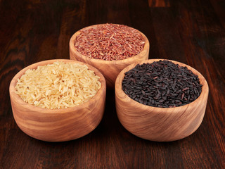 Uncooked black, red and integral rice in olive wood bowls, on a dark rustic wooden board.
