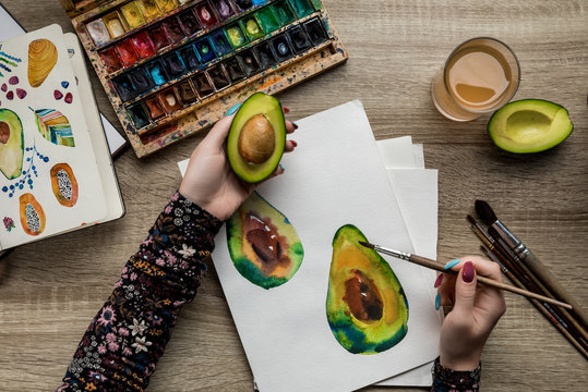 top view of female hands drawing avocado with watercolor paints and paintbrush on wooden table