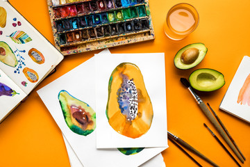 Fototapeta na wymiar top view of avocado and papaya drawings next to colored paints, paintbrushes on yellow background