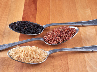 Uncooked black, red and integral rice on silver spoons, on a rustic wooden board