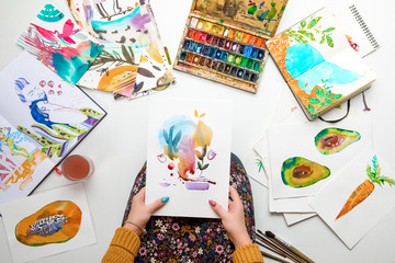 top view of woman holding watercolor drawing while surrounded by color pictures