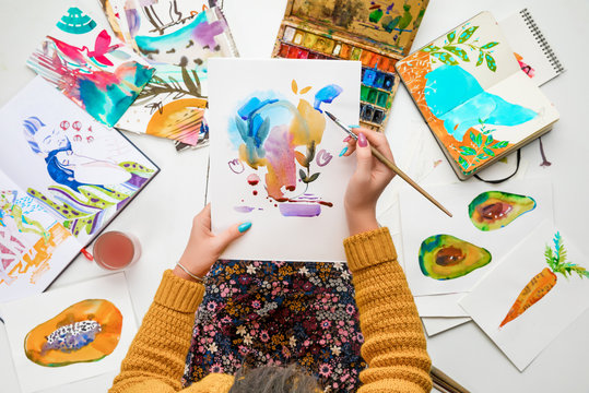 top view of woman holding drawing on knees and painting in it with watercolors paints while surrounded by colored pictures