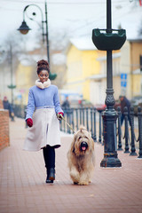 African girl in a coat walking with a dog of the Briard breed on a city street on a winter day.