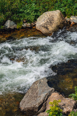 Big boulders in mountain creek close-up. Rapids of river with copy space. Fast water stream along beautiful vegetation. Fast flow near wet stones. Background of clean waves. Rich flora of highlands.